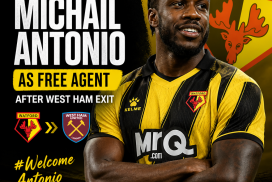 Michail Antonio signing a Watford jersey, smiling at a press conference with Watford logo in the background.